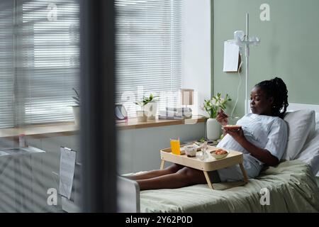 Patient dans la chambre d'hôpital avec plateau de nourriture sur le lit, appréciant le jus d'orange du petit déjeuner, des fruits, et sandwich sur le plateau tandis que le patient détend les plantes et les livres sur le rebord de la fenêtre Banque D'Images