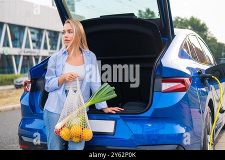 Jeune femme à l'épicerie se tient près du coffre ouvert d'une voiture électrique bleue dans un cadre urbain. Banque D'Images