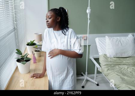 Femme enceinte debout près d'une fenêtre de l'hôpital tient son ventre et regarde dehors. Lit d'hôpital et plantes en pot en arrière-plan créant un environnement apaisant Banque D'Images