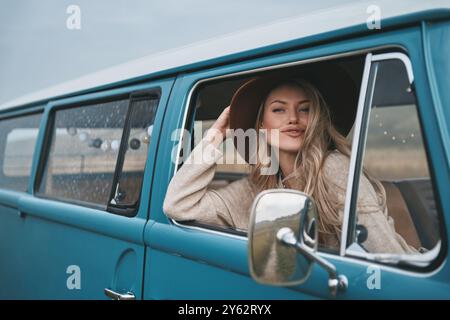 Superbe fille. Attrayant jeune femme souriante regardant par la fenêtre de la camionnette et ajustant son chapeau tout en profitant du voyage en voiture Banque D'Images