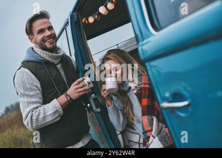 Pas de soucis et pas de précipitation. Beau jeune couple ayant des boissons chaudes et souriant tout en profitant de leur voyage sur la route Banque D'Images