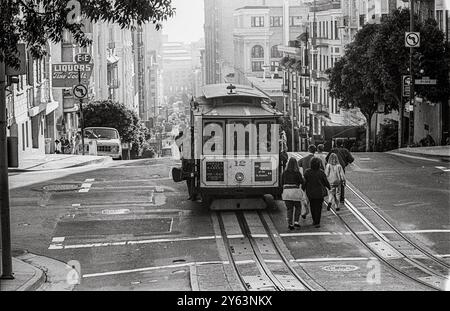 Un téléphérique fait son chemin dans une rue abrupte de San Francisco, transportant des passagers tandis que les piétons marchent le long des voies. Le mode de transport emblématique offre une expérience par excellence à San Francisco. Banque D'Images