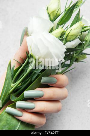 Une main avec de longs ongles élégants tient un bouquet de fleurs blanches fraîches. Les verts et les blancs doux contrastent magnifiquement avec un dos texturé léger Banque D'Images