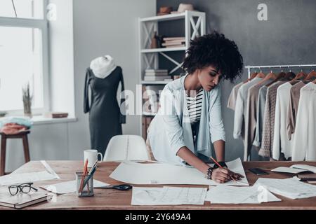 Transformer les idées en vêtements. Belle jeune femme africaine travaillant sur des croquis tout en se tenant debout dans son atelier près des vêtements suspendus sur les étagères Banque D'Images