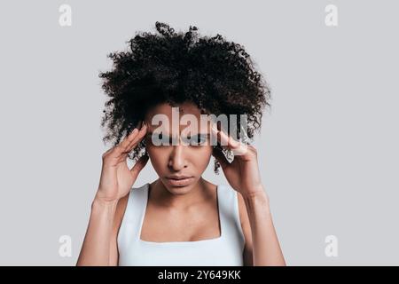 Terrible mal de tête. Frustré jeune femme africaine en vêtements décontractés touchant sa tête avec les mains et faisant le visage tout en se tenant debout sur fond gris Banque D'Images