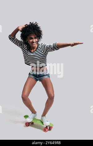 J'aime le skateboard. Plein plan studio d'une jeune femme africaine ludique en tenue décontractée faisant des gestes et souriant tout en skateboard contre le gris Banque D'Images