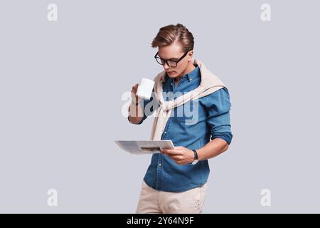 Lire les dernières nouvelles. Beau jeune homme lisant le journal et tenant une tasse tout en se tenant debout sur fond gris Banque D'Images