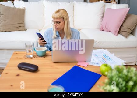 Jeune femme d'affaires tenant un smartphone tout en travaillant avec un ordinateur portable au bureau à domicile, en utilisant des applications de conception réactive multi-plateforme pour l'analyse de projet Banque D'Images