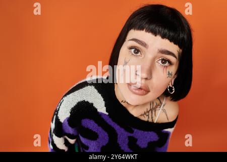 Une jeune femme présente son style unique avec des cheveux courts, des tatouages et un pull vibrant dans un cadre de studio. Banque D'Images