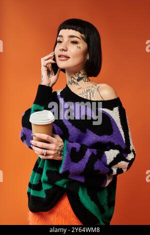 Une jeune femme élégante aux cheveux courts et aux tatouages tient une tasse à café tout en posant contre un mur orange vif. Banque D'Images