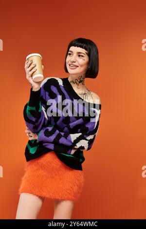 Une jeune femme aux cheveux courts et aux tatouages colorés sourit brillamment tout en tenant une tasse à café dans un cadre de studio. Banque D'Images