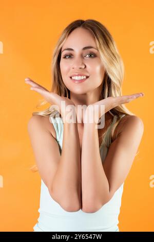 Heureuse jeune femme blonde avec coiffure décontractée posant sur fond orange et tenant les mains sous son menton Banque D'Images