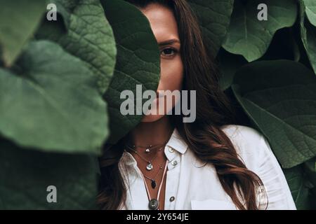 Naturellement belle. Gros plan d'une jeune femme regardant la caméra tout en se tenant parmi les feuilles à l'extérieur Banque D'Images