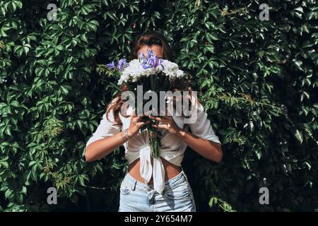 Juste être elle-même. Gros plan de jeune femme attrayante en vêtements décontractés couvrant le visage avec un bouquet de fleurs tout en se tenant devant le lierre en plein air Banque D'Images