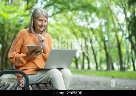 Femme mature appréciant l'apprentissage dans le parc, en utilisant un ordinateur portable et un bloc-notes pour la formation en ligne. Entourée d'une végétation luxuriante, elle s'assoit sur un banc, s'intéresse à la technologie et profite de possibilités éducatives. Banque D'Images