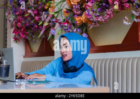 portrait de belle jeune fille asiatique portant hijab souriant en tapant candidement des données dans l'ordinateur portable tandis que casque autour du cou dans le café intérieur. pour un Banque D'Images