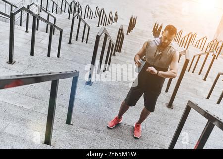 Se reposer brièvement. Beau jeune homme en vêtements de sport regardant son tracker de fitness tout en se tenant debout sur les marches à l'extérieur Banque D'Images