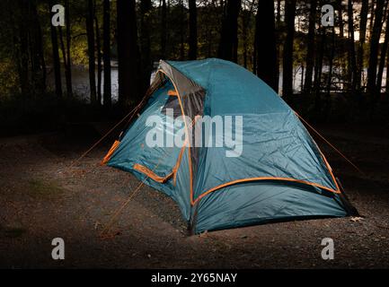 Tente bleue légère pour le sac à dos briller dans les lumières tandis que peu de lumière du soleil brille sur le lac derrière à travers la forêt épaisse soulignant la paix Banque D'Images