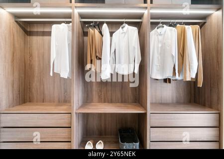 Armoire moderne et spacieuse avec une finition en bois, soigneusement organisée avec des vêtements blancs et beiges, reflétant le design intérieur sophistiqué de Banque D'Images