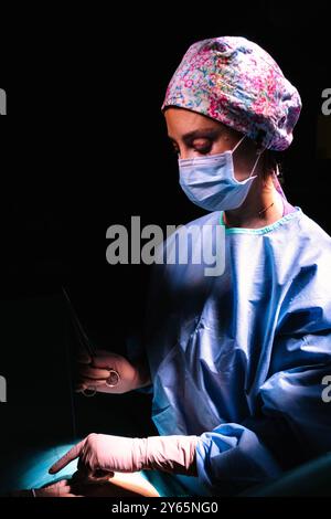 Une chirurgienne est intensément concentrée tout en effectuant une intervention chirurgicale, vêtue de gommages bleus, d'une casquette florale et d'un masque chirurgical bleu sous DRAM Banque D'Images