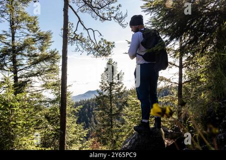 Un randonneur, portant un bonnet et portant un sac à dos, se tient sur un rocher rocheux, regardant les montagnes verdoyantes et le ciel clair de l'intérieur d'un ombragé Banque D'Images