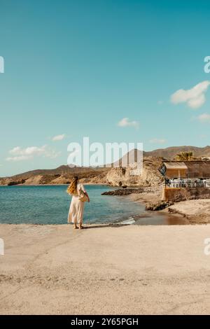 Vue arrière d'une femme méconnaissable dans une robe blanche regardant la mer à Isleta del Moro, Cabo de Gata, capturant un sce balnéaire serein et pittoresque Banque D'Images