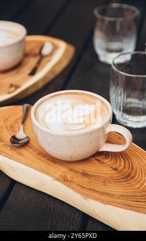 Vue de dessus d'un cappuccino fraîchement infusé avec une présentation artistique en mousse, servi sur une planche de bois incurvée à côté d'un verre d'eau claire et un autre coff Banque D'Images