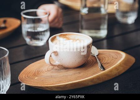 Vue de dessus d'un cappuccino fraîchement infusé dans une tasse en céramique mouchetée, servi sur une soucoupe en bois avec une cuillère en métal, dans un café. Banque D'Images