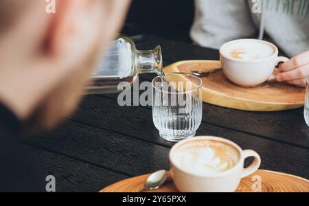 Une personne rognée méconnaissable verse de l'eau d'une bouteille dans un verre clair à une table de café en bois sombre, accompagnée d'un cappucc magnifiquement présenté Banque D'Images