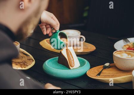 Homme taillé méconnaissable versant de la sauce caramel sur un cheesecake dans un cadre de café. Une sélection de boissons, y compris du café, est également visible sur le Banque D'Images