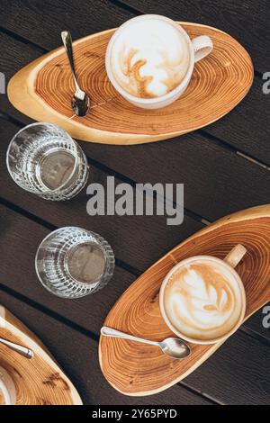 Vue de dessus de deux cappuccinos aux motifs en mousse artistiques, servis sur des assiettes en bois à côté de verres d'eau clairs, exposés sur une table en bois sombre. Banque D'Images
