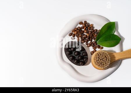 Un éventail de grains de café et un gommage naturel au café affiché sur un plat en céramique blanche, accompagné de feuilles vertes fraîches et d'une brosse en bois idéale pour Banque D'Images