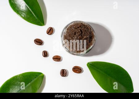 Un gommage au café maison frais affiché dans un pot en verre transparent entouré de grains de café et de feuilles vertes sur un fond blanc, soulignant son natur Banque D'Images