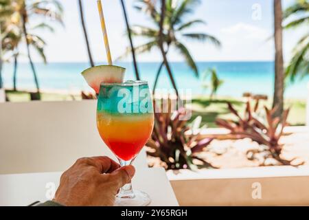 Une vue rapprochée d'un cocktail tropical coloré, avec une tranche de pastèque comme ornement en arrière-plan, les sables immaculés et les palmiers Banque D'Images