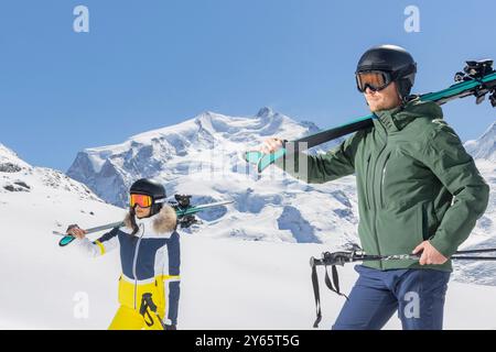 Un homme et une femme vêtus d'une tenue de ski d'hiver à la mode contre un paysage montagneux enneigé saisissant, portant des skis et des bâtons, incarnant la vie active Banque D'Images