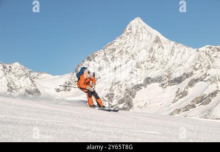 Un skieur vêtu d'un équipement orange vif sculpte à travers la poudre fraîche sur une vaste pente ouverte avec des pics dentelés et enneigés s'élevant en arrière-plan sous un Clea Banque D'Images