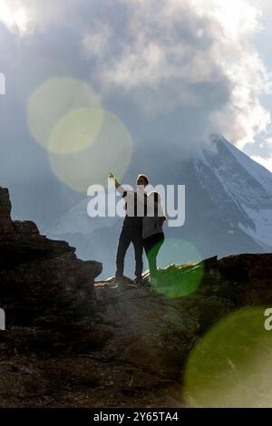 Couple embrassant et pointant tout tout tout en se tenant debout au sommet rocheux, avec un pic de montagne enneigé sur le fond Banque D'Images