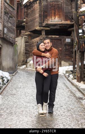 Couple asiatique profitant de vacances de Noël, s'embrassant chaleureusement dans une rue pavée de Zermatt, avec des maisons en bois rustiques en arrière-plan Banque D'Images