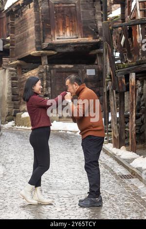 Couple asiatique profite d'un moment tendre dans une rue enneigée de Zermatt, avec l'homme embrassant la main de la femme. Les deux sont souriants et habillés en hiver décontracté Banque D'Images