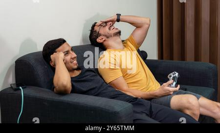 Un couple gay partage un moment léger tout en jouant à des jeux vidéo sur un canapé, mettant en évidence un charmant scénario LGBTQIA+ à domicile qu'ils aiment rire Banque D'Images