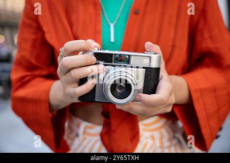 Gros plan des mains d'une personne tenant un appareil photo argenté à l'ancienne, contrastant avec leur veste orange vif et leur collier vert. Banque D'Images