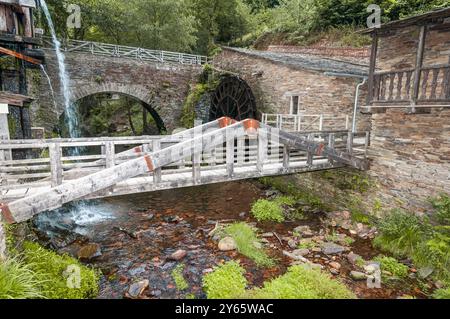 Une vue panoramique sur un ancien moulin à eau avec une grande roue à eau, un pont en pierre adjacent et une cascade, entouré d'une végétation luxuriante Banque D'Images