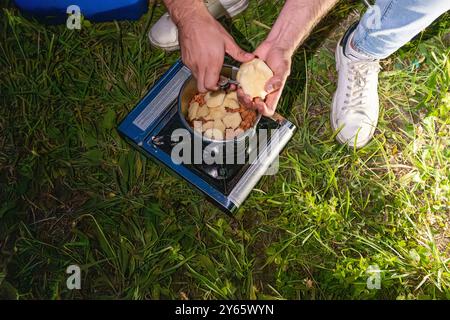 D'en haut, un campeur mâle méconnaissable et cultivé prépare un repas sur un poêle portable fixé sur l'herbe, avec des mains tenant des pommes de terre tranchées au-dessus d'un pot Banque D'Images