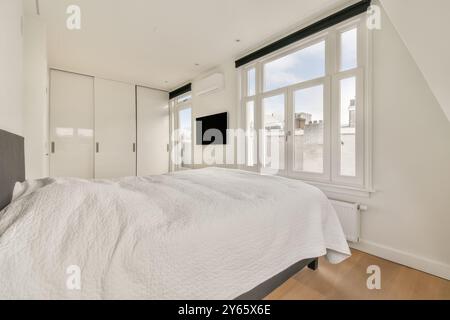 Cette chambre minimaliste lumineuse et aérée dispose d'un grand lit, d'élégants meubles blancs, de planchers de bois franc et d'une grande lumière naturelle provenant de grandes fenêtres. Banque D'Images