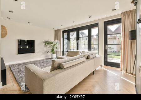 Un salon spacieux et moderne avec de grandes fenêtres, un mobilier élégant et un confortable canapé beige baigné de lumière naturelle. Banque D'Images