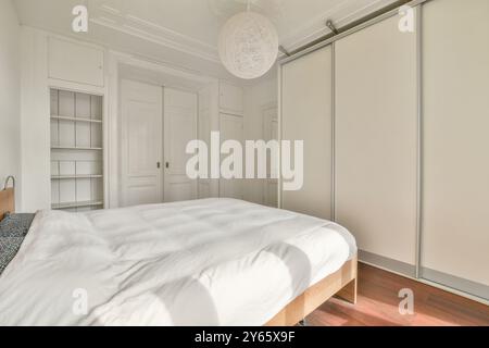 Un intérieur de chambre lumineux et aéré avec des lignes épurées, un grand lit avec une literie blanche, des armoires blanches intégrées et des moulures classiques. Banque D'Images