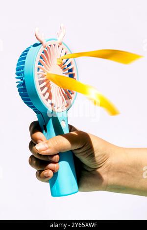 Une image en gros plan capturant un ventilateur électrique bleu portable, activement tourné par une main sur un fond clair les rubans jaunes flottent de manière vibrante à partir de t Banque D'Images