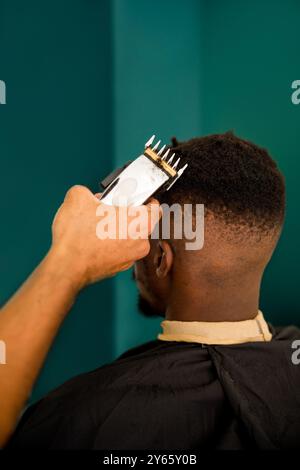 Un barbier professionnel coupe les cheveux d'un jeune homme noir dans un salon de coiffure côtier stylisé, offrant un look propre et moderne sur fond sarcelle Banque D'Images