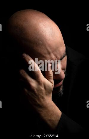 Un gros plan d'un homme chauve en détresse couvrant une partie de son visage et de son front avec sa main il semble être dans la tristesse ou la douleur, placé contre un noir sombre Banque D'Images