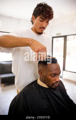 Un barbier habile coupe soigneusement les cheveux d'un homme noir avec des ciseaux dans un salon de coiffure moderne et lumineux, assurant une attention méticuleuse aux détails Banque D'Images